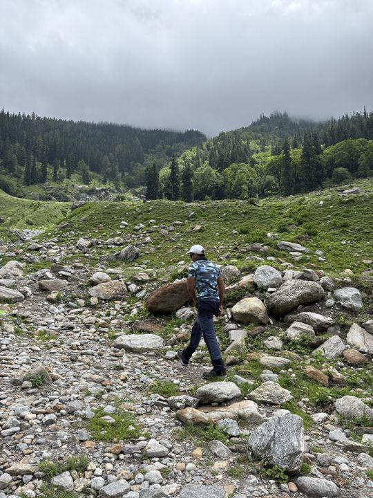 A trekker in a patterned shirt walking across boulders below the forest