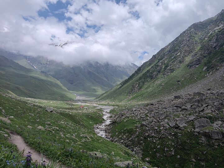 A trail through wildflowers above a stream, tents on the valley floor
