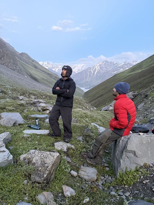 Two trekkers at a rocky camp, snow peaks at the valley head
