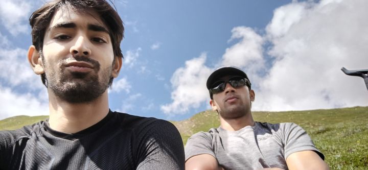 A low-angle selfie of two friends against blue sky and a green slope