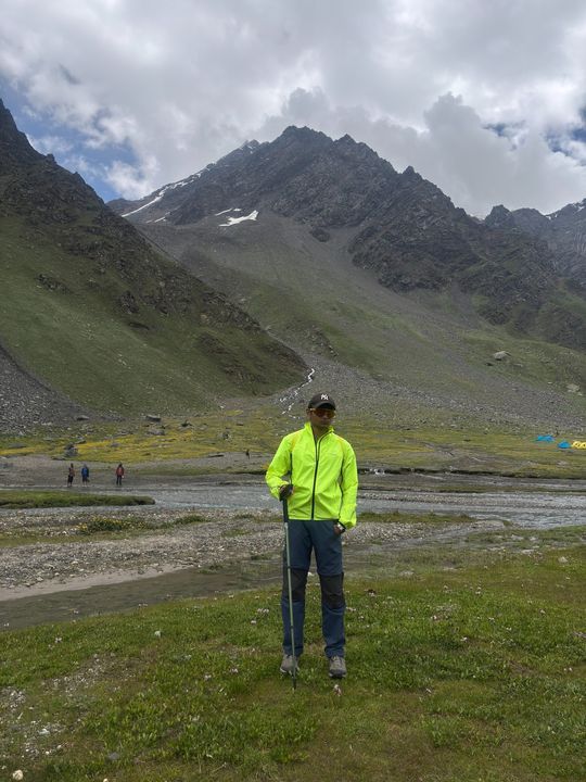 A trekker in a neon jacket on a meadow by a braided river, tents behind