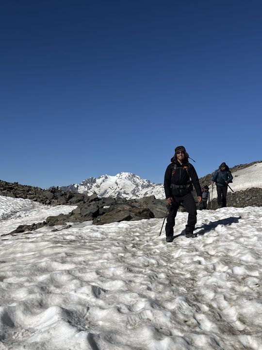A trekker in a fur hat on the snow below a white peak, others following