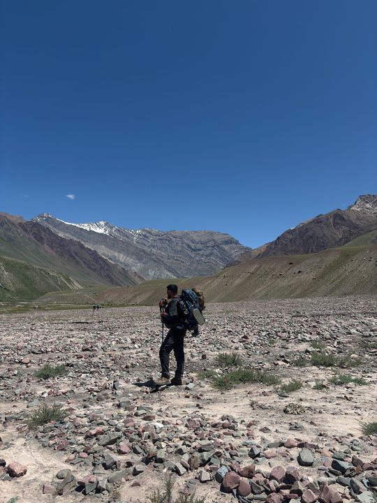 A trekker with a pack on a pebbled plain below banded mountains